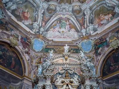 The image shows the ornate, illusionistic ceiling and upper facade of a Baroque church interior, featuring frescoes, stucco sculptures, and a trompe-l'œil architectural design.