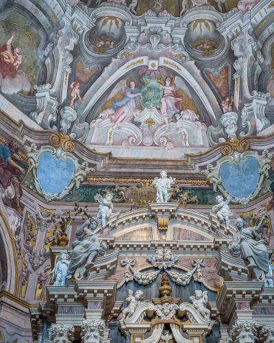 The image shows the ornate, illusionistic ceiling and upper facade of a Baroque church interior, featuring frescoes, stucco sculptures, and a trompe-l'œil architectural design.