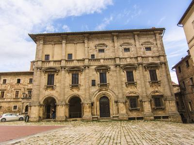 A low-angle photograph of a large, three-story Mannerist stone palace with rusticated masonry, arched ground-floor loggias, and a prominent central entrance, situated on a cobblestone piazza under a blue sky.