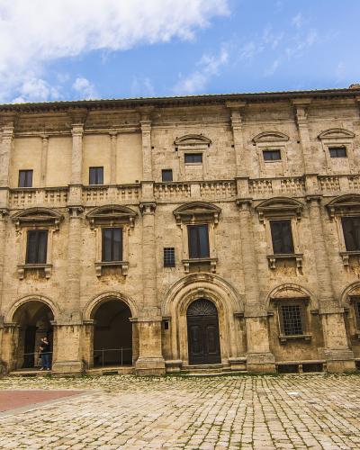 A low-angle photograph of a large, three-story Mannerist stone palace with rusticated masonry, arched ground-floor loggias, and a prominent central entrance, situated on a cobblestone piazza under a blue sky.