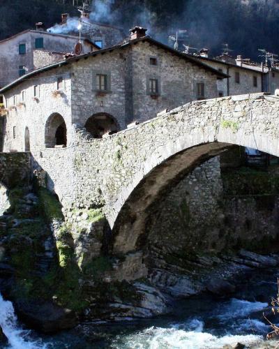 A stone arch bridge spanning a rushing river with a waterfall, backed by a large stone building with arched openings and residential structures on a hillside.