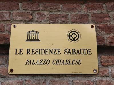 A brass plaque mounted on a red brick wall, displaying the UNESCO logo, the text "LE RESIDENZE SABAUDE," and "PALAZZO CHIABLESE," along with a small inscription at the bottom reading "TORINO."