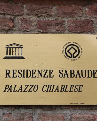 A brass plaque mounted on a red brick wall, displaying the UNESCO logo, the text "LE RESIDENZE SABAUDE," and "PALAZZO CHIABLESE," along with a small inscription at the bottom reading "TORINO."