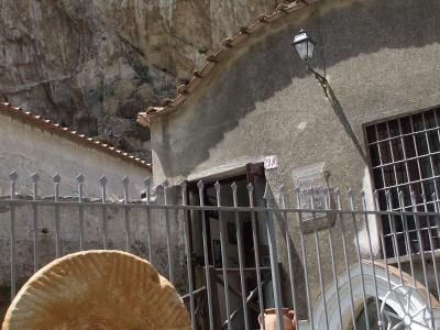 A stone building with a curved facade and terracotta roof tiles, situated at the base of a rocky cliff, featuring a large millstone in the foreground and a plaque on the wall.