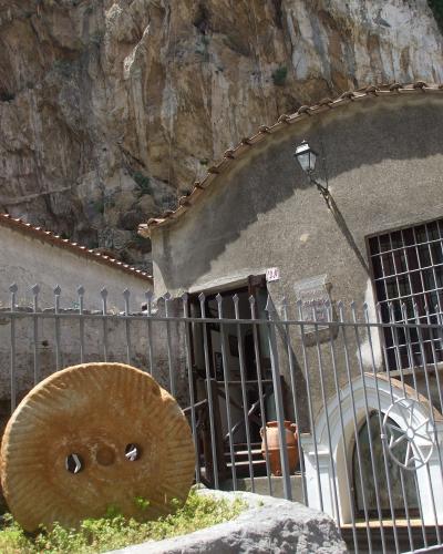 A stone building with a curved facade and terracotta roof tiles, situated at the base of a rocky cliff, featuring a large millstone in the foreground and a plaque on the wall.