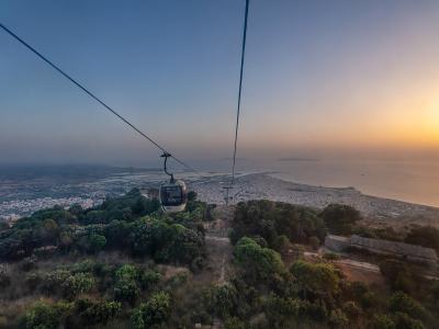 A cable car gondola suspended on cables above a hillside with trees, overlooking a coastal city and sea at sunset; no visible signage or text in the image itself.