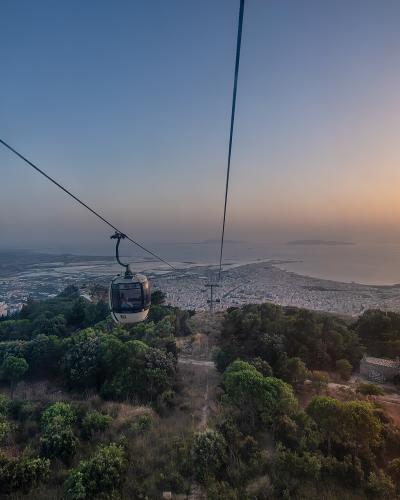 A cable car gondola suspended on cables above a hillside with trees, overlooking a coastal city and sea at sunset; no visible signage or text in the image itself.