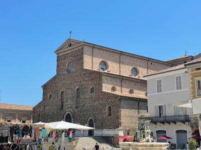 A large, historic brick cathedral with a stepped facade, round and arched windows, and a cross on the roof, situated in a public square with market stalls, a fountain, and adjacent buildings under a clear blue sky.