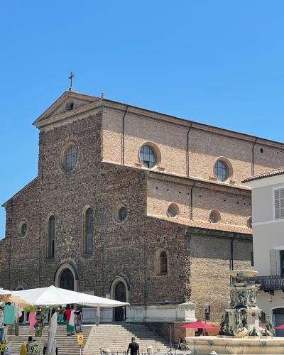 A large, historic brick cathedral with a stepped facade, round and arched windows, and a cross on the roof, situated in a public square with market stalls, a fountain, and adjacent buildings under a clear blue sky.