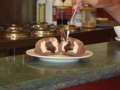 A close-up shot of a chocolate-coated ice cream sphere cut in half to reveal a creamy custard center, being served with a spoon on a white plate atop a green marble counter, with condiment bottles and metal lids visible in the background.