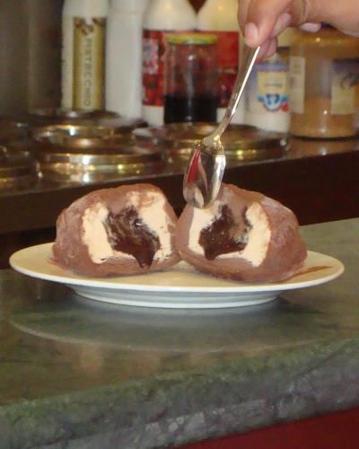 A close-up shot of a chocolate-coated ice cream sphere cut in half to reveal a creamy custard center, being served with a spoon on a white plate atop a green marble counter, with condiment bottles and metal lids visible in the background.
