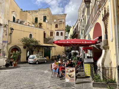 A sunlit cobblestone street in a historic Italian village, featuring yellow stucco buildings, a red "Lounge Bar Dolcezza & Bontà" umbrella, outdoor cafe seating, a silver car, and a small religious shrine on the right.