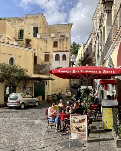 A sunlit cobblestone street in a historic Italian village, featuring yellow stucco buildings, a red "Lounge Bar Dolcezza & Bontà" umbrella, outdoor cafe seating, a silver car, and a small religious shrine on the right.