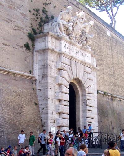 The image shows a stone entrance gate set into a high brick wall, featuring a sculpted coat of arms and the inscription "MUSEI VATICANI" above the arch, with tourists queuing behind metal barriers.