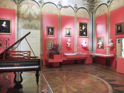 The image shows an interior museum gallery with pink walls, a harpsichord in the foreground, display cases, portraits, and busts, under a frescoed ceiling.