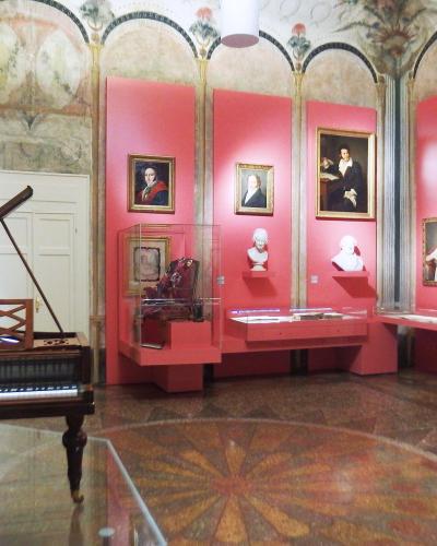 The image shows an interior museum gallery with pink walls, a harpsichord in the foreground, display cases, portraits, and busts, under a frescoed ceiling.