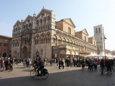 The image shows the Ferrara Cathedral (Cattedrale di San Giorgio) in Ferrara, Italy, identifiable by its distinctive striped facade, Romanesque-Gothic architecture, and adjacent bell tower, situated in a large public square with people and bicycle