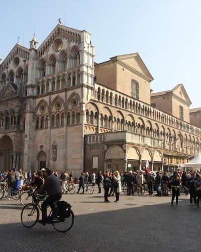 The image shows the Ferrara Cathedral (Cattedrale di San Giorgio) in Ferrara, Italy, identifiable by its distinctive striped facade, Romanesque-Gothic architecture, and adjacent bell tower, situated in a large public square with people and bicycle
