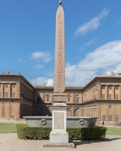 A tall Egyptian-style obelisk with hieroglyphs stands on a pedestal in front of a large, rusticated Renaissance palace building under a blue sky.