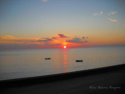 A sunset over a calm sea with two small boats floating near the horizon, viewed from a dark concrete promenade or seawall in the foreground; watermark reads "Foto Melania Maquino".