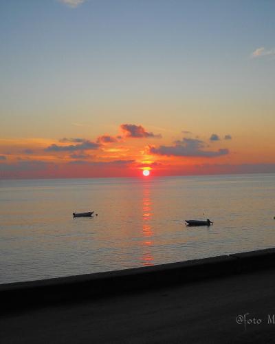 A sunset over a calm sea with two small boats floating near the horizon, viewed from a dark concrete promenade or seawall in the foreground; watermark reads "Foto Melania Maquino".