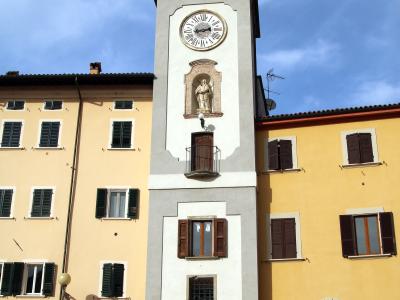 A tall, slender clock tower with a white and grey facade, a large clock face, a statue niche, and a domed roof, situated between two yellow buildings in a European town square with parked cars and a "SALE e TABACCHI" sign.