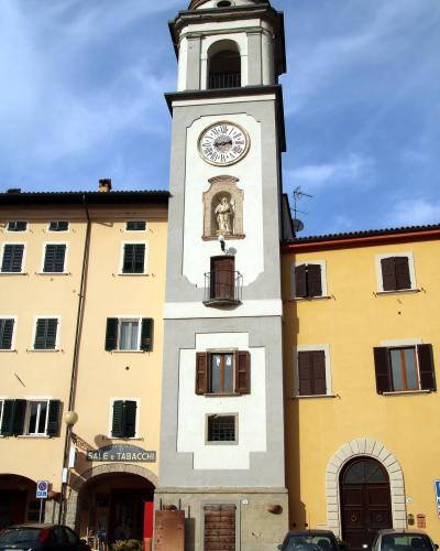 A tall, slender clock tower with a white and grey facade, a large clock face, a statue niche, and a domed roof, situated between two yellow buildings in a European town square with parked cars and a "SALE e TABACCHI" sign.