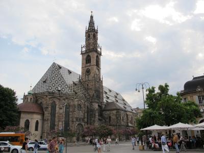 A large Gothic-style cathedral with a distinctive patterned roof and tall ornate spire, situated in a public square with pedestrians, outdoor café umbrellas, and vehicles nearby.