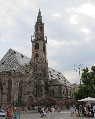 A large Gothic-style cathedral with a distinctive patterned roof and tall ornate spire, situated in a public square with pedestrians, outdoor café umbrellas, and vehicles nearby.