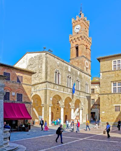 A sunlit Renaissance-era town square with a prominent brick clock tower, arched loggia, stone buildings, and pedestrians walking on patterned paving; a red awning and EU flag are visible.