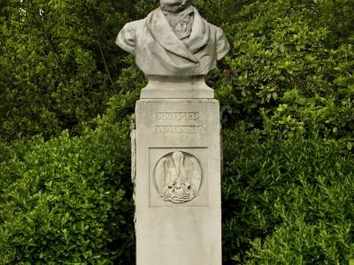 A stone bust of Richard Wagner on a pedestal with his name inscribed, set against a backdrop of dense green hedges and trees.