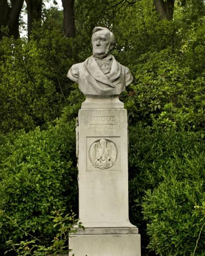 A stone bust of Richard Wagner on a pedestal with his name inscribed, set against a backdrop of dense green hedges and trees.