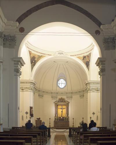 The image shows the interior of a church with a central nave, wooden pews, a domed apse, and an altar featuring a crucifix, with Latin inscriptions on the arch above the dome.