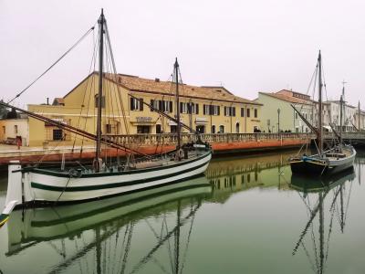 A canal scene featuring traditional wooden sailing boats (barche da pesca) moored along a quay with yellow buildings in the background, under an overcast sky.