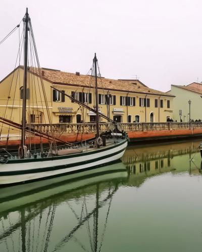 A canal scene featuring traditional wooden sailing boats (barche da pesca) moored along a quay with yellow buildings in the background, under an overcast sky.