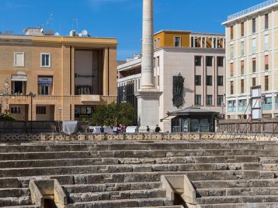 The image shows the partially excavated stone seating tiers of an ancient Roman amphitheater in the foreground, with a tall column topped by a statue (the Column of Saint Oronzo) and surrounding modern buildings in the background under a blue sky.