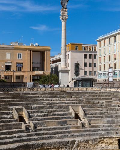 The image shows the partially excavated stone seating tiers of an ancient Roman amphitheater in the foreground, with a tall column topped by a statue (the Column of Saint Oronzo) and surrounding modern buildings in the background under a blue sky.