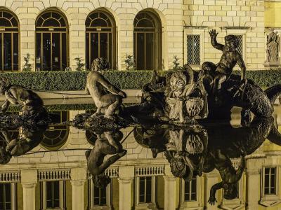 A nighttime view of a large, ornate fountain featuring mythological figures and a central sea-monster sculpture, with its reflection visible in the water, set against the illuminated neoclassical facade of a building with arched windows.