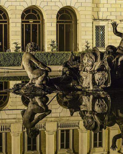 A nighttime view of a large, ornate fountain featuring mythological figures and a central sea-monster sculpture, with its reflection visible in the water, set against the illuminated neoclassical facade of a building with arched windows.