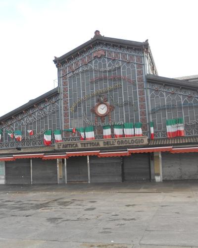 The image shows the "L'antica tettoia dell'orologio" (The old clock canopy), a historic iron-and-glass market hall in Turin, Italy, decorated with Italian flags and signage referencing the 150th anniversary of Italian unification.