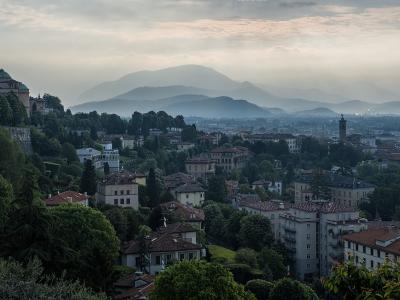 A misty, elevated landscape view of a town with terracotta-roofed buildings nestled among green hills, with a prominent domed building on the left and layered mountains in the background under a cloudy sky.