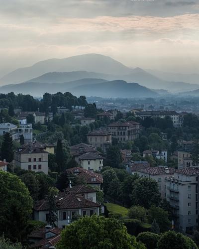 A misty, elevated landscape view of a town with terracotta-roofed buildings nestled among green hills, with a prominent domed building on the left and layered mountains in the background under a cloudy sky.