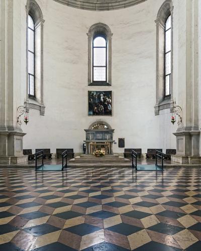The image shows the interior of a chapel with a distinctive black, white, and tan checkered floor, large Baroque-style paintings on the walls, wooden confessionals, and an altar at the far end under a dome.