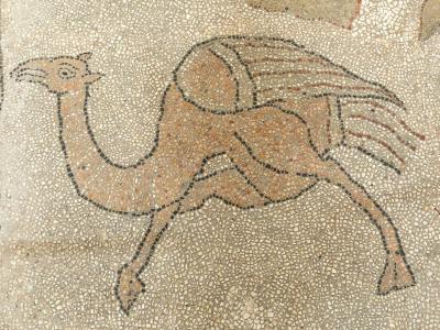 A close-up photograph of a medieval floor mosaic depicting a camel-like creature with wings, rendered in small tesserae of beige, brown, and black stone.