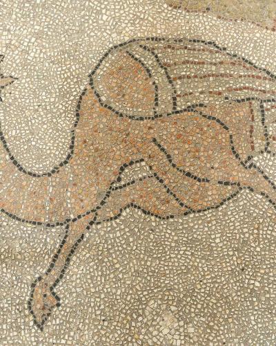 A close-up photograph of a medieval floor mosaic depicting a camel-like creature with wings, rendered in small tesserae of beige, brown, and black stone.