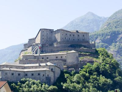 A large, multi-tiered stone fortress built into a steep, forested hillside with mountains in the background; no visible signage or text is present in the image.