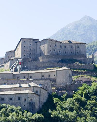 A large, multi-tiered stone fortress built into a steep, forested hillside with mountains in the background; no visible signage or text is present in the image.