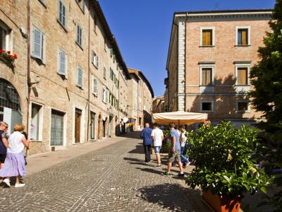 A sunlit, cobblestone pedestrian street in an Italian historic town, flanked by multi-story brick buildings with shuttered windows and arched doorways, featuring pedestrians and a street vendor's umbrella.