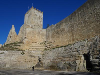 A low-angle shot of a large medieval stone fortress built atop a steep, layered rock cliff under a clear blue sky, with a statue and a person visible at the base.
