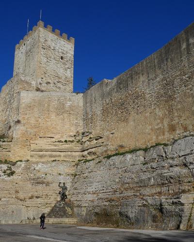 A low-angle shot of a large medieval stone fortress built atop a steep, layered rock cliff under a clear blue sky, with a statue and a person visible at the base.